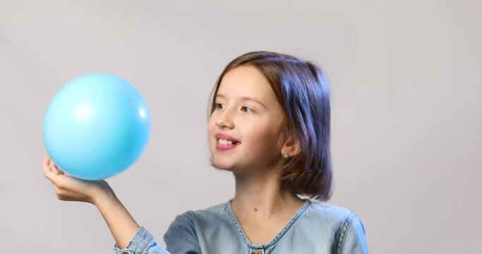 Pretty little girl in casual denim dress blowing, inflate blue balloon on white background, studio