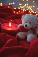 White teddy bear and burning candle on red fabric with bokeh background