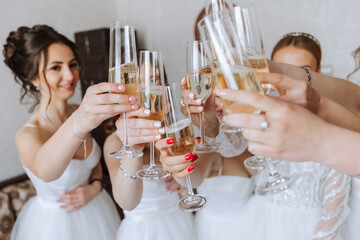 Bride with merry girlfriends at the wedding drink champagne from glasses. Bride and girlfriends hug in the room. Morning Bride and girlfriends. White dress.