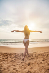 Fototapeta na wymiar Back view young woman in white swimsuit going step with hands to side at ocean background, summertime. Relaxing and enjoyable vacation on tropical sea. Travel vacation holiday concept. Copy text space