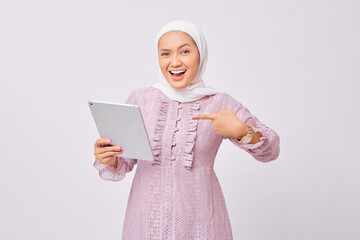 Portrait of smiling beautiful young Asian Muslim woman wearing hijab and purple dress pointing finger at digital tablet isolated on white studio background