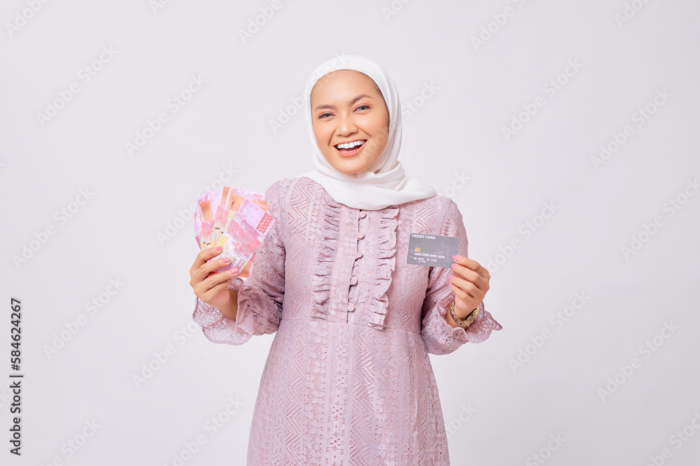 Wall mural Cheerful young Asian Muslim woman wearing hijab and purple dress holding credit card and money banknotes isolated on white studio background