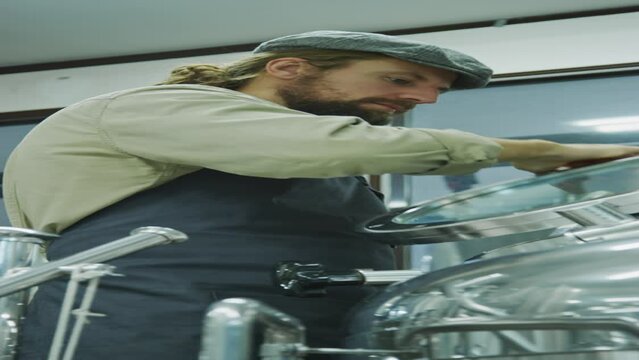 Waist-up vertical shot of young Caucasian male microbrewery owner in cap and apron working alone on brewhouse system and opening hatch of mash tun with sight glass