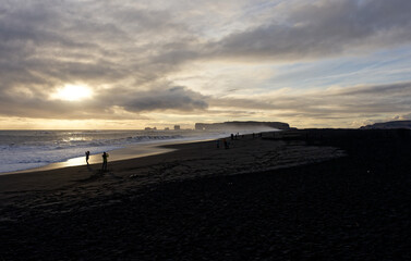 coucher de soleil au bord d'une page de salble noir avec des personne en ombre chinoise donnant sur les falaises abtutes de Dyrholaey en Islande 
