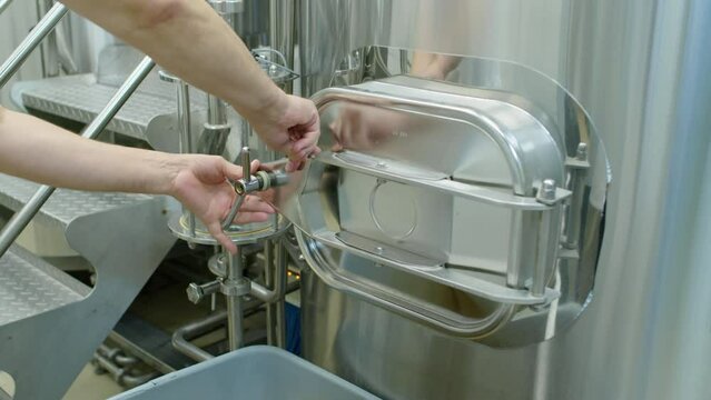 Close-up shot of hands of anonymous brewery worker twisting lock and opening hatch on large stainless steel lauter tun, preparing to remove spent grain