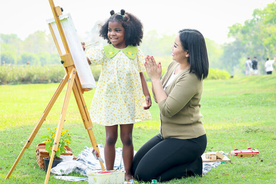 Cute smile Asian African girl child with black curly hair painting on canvas, mother applauded in garden outdoors, kid and parent draw picture together in summer park, mom and daughter happy family.
