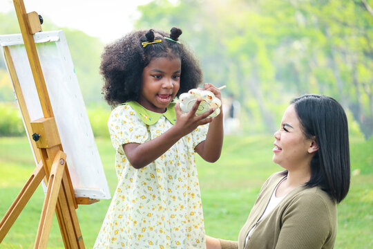 Cute smiling Asian African girl child with black curly hair painting on canvas with mother green garden outdoor, kid and parent draw picture together in summer park, mom and daughter happy family.