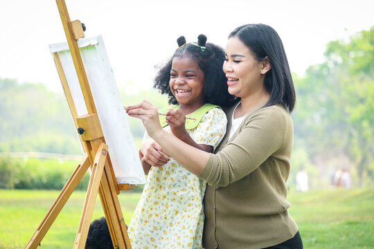 Cute smiling Asian African girl child with black curly hair painting on canvas with mother green garden outdoor, kid and parent draw picture together in summer park, mom and daughter happy family.