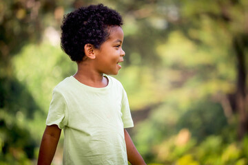 Portrait of happy smiling African boy with black curly hair standing and laughing outdoor with blurred green garden background, beautiful kid play outside park on sunny day, cute child play in summer.