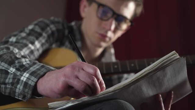 a young man with glasses writes the text of a song with an acoustic guitar in a cozy room.
