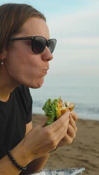 A young woman on the seashore, with her mouth wide open, bites off a large piece of burger. Picnic by the sea. Vertical video