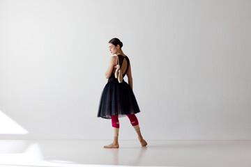 Side view. Young graceful female ballet dancer or classic ballerina wearing black dress dancing over white background © Lustre Art Group 