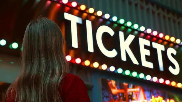 Smiling young woman looking at the vintage carnival tickets sign at the amusement park, bright concept of fun. Family weekend. Girl at an evening fanfare against a backdrop of colorful lights.
