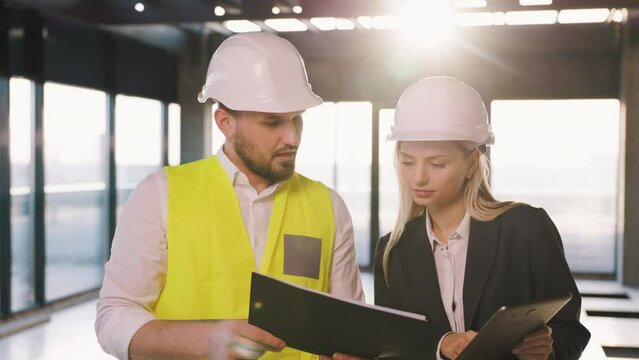 A civil engineer and an architect manager are discussing the plan for the construction of a business center. The workers are wearing helmets. Sun rays are shining through the window