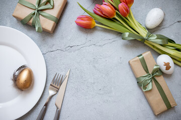 Fototapeta na wymiar a white dinner plate, a bouquet of tulips, gifts, cutlery and easter eggs on a gray concrete stylish background. copy space. flat lay. top view. place for text