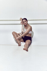 Adorable ballerina leaning on dance bar and resting after hard work in dance hall over white background © Lustre Art Group 