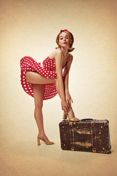 Oops. Sensual, cute, beautiful blonde girl in attractive red dress posing with suitcase against brown background. Wind blowing. Travelling. Concept of retro fashion, beauty, 50s, 60s. Pin-up style
