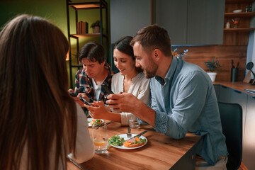 Sitting by the table with smartphones in hands. Group of people are on the kitchen, having dinner, spending time together