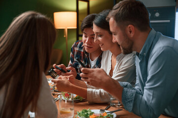 Fototapeta premium Sitting by the table with smartphones in hands. Group of people are on the kitchen, having dinner, spending time together
