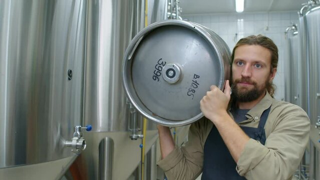 Waist-up tracking shot of young Caucasian male brewery employee walking along large brite tanks in fermentation room and carrying heavy beer keg on shoulder