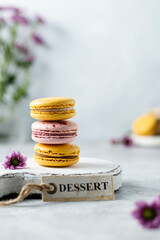 Obraz na płótnie Canvas Stack of yellow and pink colored macaron cookies. French mini dessert with almond flour. Violet flowers on grey background. Spring or summer mood. Tag dessert on a table. Copy space