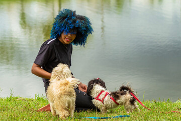 Uma jovem negra com cabelos tingidos de azul está sentada no gramado ao lado de um lago com seus cachorros.