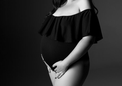 A pregnant woman in a black bodysuit on a gray background with a place for text. Belly of a pregnant woman in close-up. Studio pregnancy photo shoot. Black and white photo. Woman in black underwear.