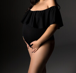 A pregnant woman in a black bodysuit on a gray background with a place for text. Belly of a pregnant woman in close-up. Studio pregnancy shooting.  Woman in black underwear