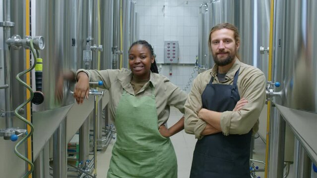 Medium wide portrait shot of two multiethnic male and female brewery employees standing in beer fermentation facility next to shiny large vats, smiling happily and looking at camera