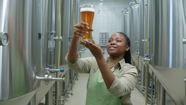 Medium arc shot of African American female brewery technologist pouring glass of freshly brewed lager from fermenter tank, lifting up, checking texture and carbonation, and smiling with satisfaction
