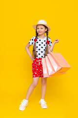 A little girl holds shopping bags. A child in a summer hat and shorts is engaged in children's shopping. A happy child in full growth on a yellow isolated background.