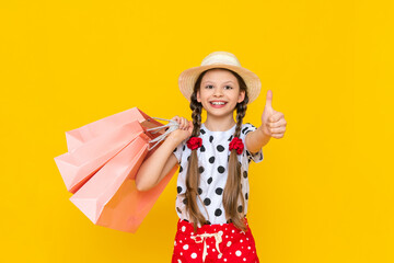 A child with paper bags from the store. Summer shopping for children. A beautiful little girl in a hat is smiling broadly.   Yellow isolated background.