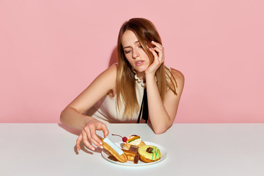 Adorable girl sitting at table and taking ecler with sad facial expression over pink background