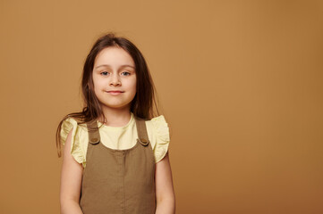Lovely Caucasian toddler girl with beautiful long hair, dressed in stylish khaki dress and yellow t-shirt, smiling at camera, isolated on beige background with copy advertising space. Children. Kids
