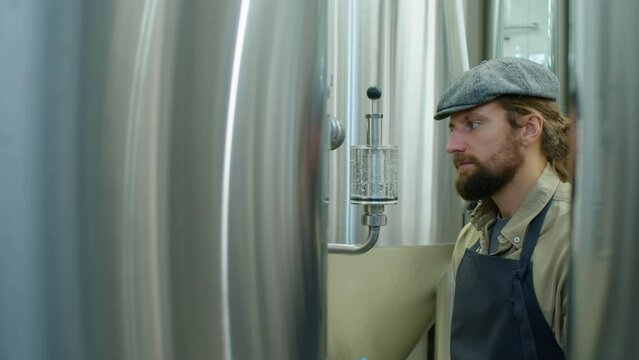 Medium shot of young Caucasian male brewery technician walking between large stainless steel fermentation tanks, stopping, tapping on pressure gauge and gas filter, and leaving
