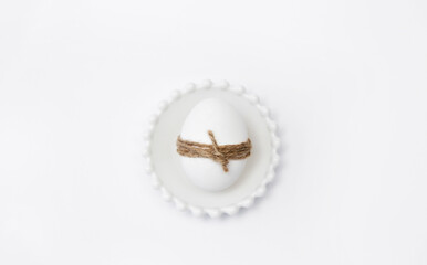 Easter holiday. A white egg tied with thread lies on plate on white background. 