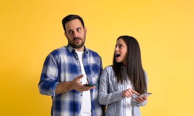 Surprised girlfriend with mouth open using mobile phone and looking at rude boyfriend rolling his eyes in irritation and ignoring her on isolated yellow background