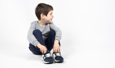 Little boy in a gray sweatshirt, blue sweatpants and sneakers on a white background sits and looks...