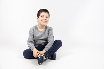 Little boy in a gray sweatshirt, blue sweatpants and sneakers on a white background sits and smiles, space for text