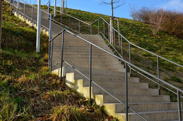stairs in a steep grassy zig zag slope will shorten and reduce the steepness. urban architecture with gray tubular railings. meadow and newly planted trees on slope. it is not wheelchair accessible