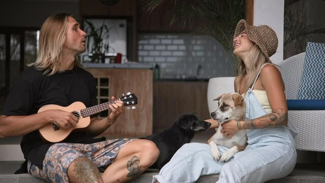 Loving family sing fun song. Happy talent man play ukulele guitar. Joyful people care cute street dogs. Lovers couple rest together. Hippies romantic date. Pets sit porch. Guitarist concert performing