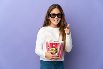 Child over isolated purple background with 3d glasses and holding a big bucket of popcorns while pointing front