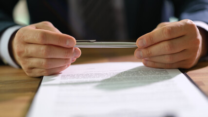 Businessman holding ballpoint pen in hands and studying information on document before signing at work in office closeup