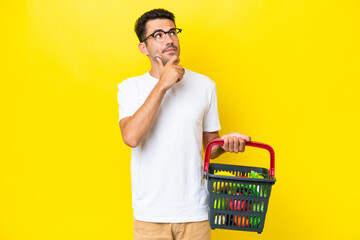 Young handsome man holding a shopping basket full of food over isolated yellow background having...