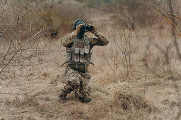 a soldier in camouflage holds binoculars. a soldier in the steppe watches the enemy