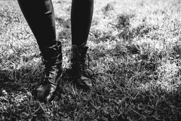 Style in park: details of feet and legs of person in black eco leather pants and black military fashion boots over grass (in black and white)