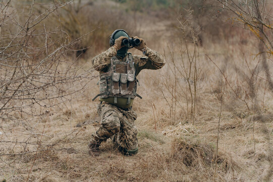 a soldier in camouflage holds binoculars. a soldier in the steppe watches the enemy