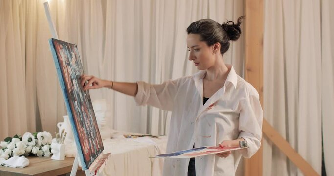 Young brunette woman artist painting picture on canvas in art studio. On the background is a windows with white curtains. The concept of art, creativity.