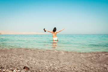 Young carefree Caucasian woman tourist dance on the beach in turquoise water alone with hands up hold slippers on summer vacation beach holidays. Solo travel adventure wanderlust