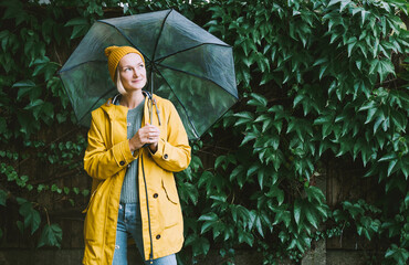 Happy woman wearing yellow raincoat holding transparent umbrella green leaves wall. Thoughtful young girl smiling outdoors at autumn season. - obrazy, fototapety, plakaty